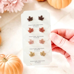 May include: Four pairs of fall-themed stud earrings with a maple leaf design. The earrings are made of glitter and come in four different colors: brown, pink, gold, and a mix of brown and gold. The earrings are displayed on white cards with the text "Couture Designs Studio" printed on them.