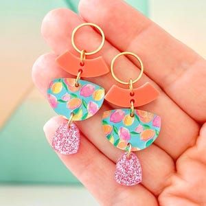 May include: A pair of colorful earrings with gold-tone hoops. The earrings feature a coral-colored arch, a turquoise semi-circle with a lemon pattern, and a pink glitter teardrop. The earrings are held in a hand.