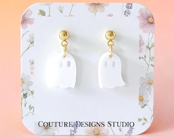 May include: A pair of white ghost-shaped dangle earrings with gold-tone ball studs. The earrings are displayed on a white card featuring a floral border and the text Couture Designs Studio printed in black at the bottom of the display card.