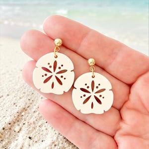 May include: A pair of cream-colored sand dollar earrings with gold-colored posts and accents. The earrings have a circular shape with a cut-out starburst design. The background shows a beach scene with sand and ocean.