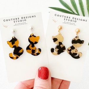 May include: Two pairs of cat-shaped earrings. One pair features a silver post and a black and orange tortoiseshell pattern. The other pair has a gold post and a black and tan tortoiseshell pattern. Each earring is on a white card with the text "COUTURE DESIGNS STUDIO".