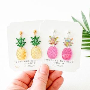 May include: Two pairs of pineapple earrings on white card stock. The earrings are made of clear acrylic with a glitter finish. One pair is yellow with green pineapple tops and the other pair is pink with multi-colored pineapple tops. The card stock has the text "Couture Designs Studio" printed on it.