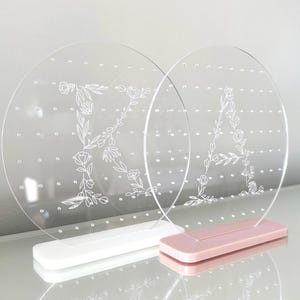 May include: Two clear acrylic earring holders with floral letter designs. One has a white base and the letter "K", the other has a pink base and the letter "A". Each holder has multiple small holes for earrings.