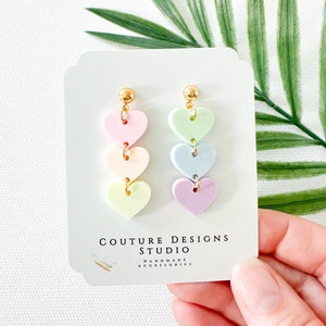 May include: Pastel pink, light blue, light green, and lavender heart shaped dangle earrings with gold accents. The earrings are on a white card with the text "Couture Designs Studio Handmade Accessories".