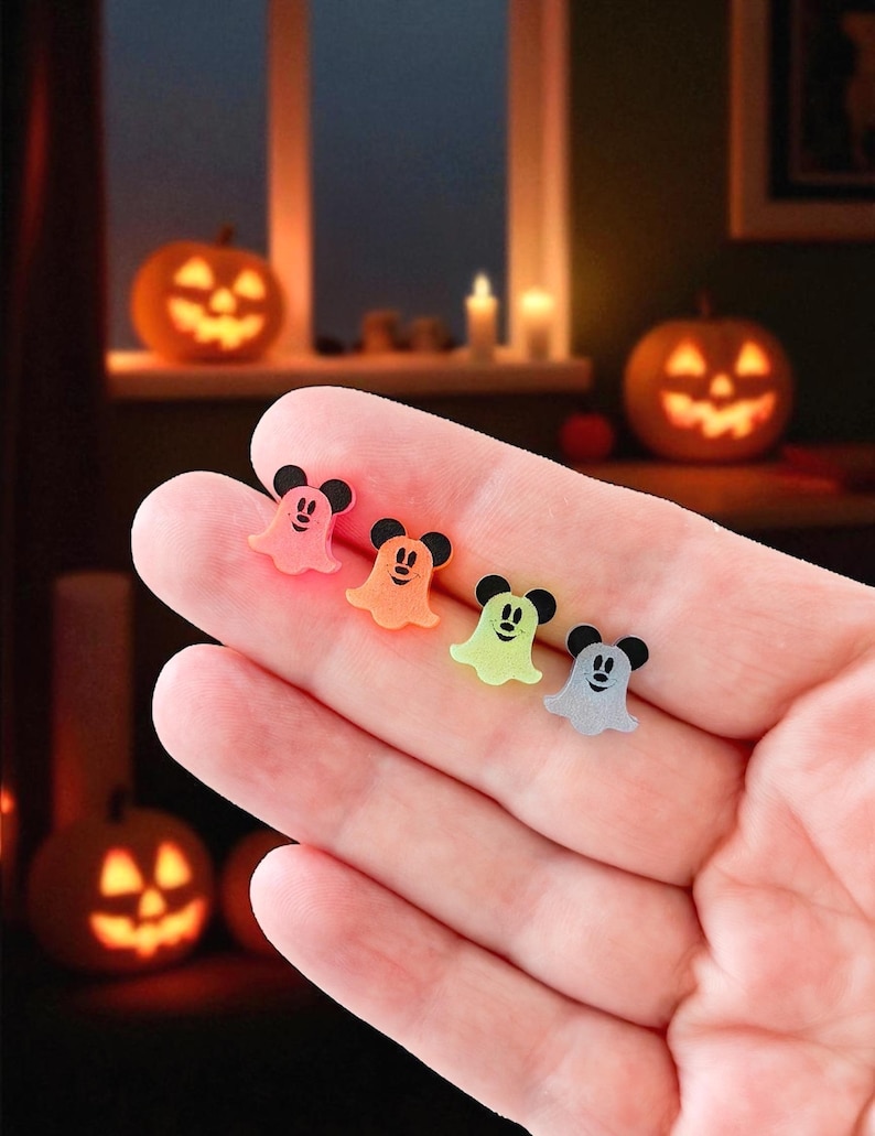 Puede incluir: Cuatro decoraciones de Halloween con forma de fantasma de colores con detalles negros de orejas de rat&oacute;n se sostienen en una mano. Los fantasmas son rosas, naranjas, verdes y grises. El fondo incluye calabazas iluminadas y una ventana.