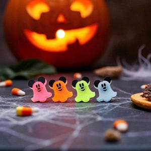 Puede incluir: Una imagen con tem&aacute;tica de Halloween con cuatro figuras coloridas en forma de fantasma con orejas de rat&oacute;n. Los fantasmas son rosas, naranjas, verdes y azules, dispuestos en fila. Una calabaza tallada con una cara brillante en el fondo.