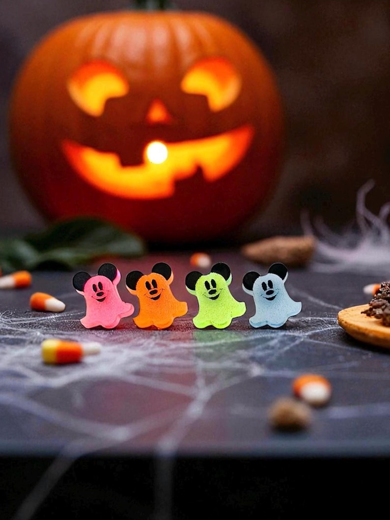 Puede incluir: Una imagen con tem&aacute;tica de Halloween con cuatro figuras coloridas en forma de fantasma con orejas de rat&oacute;n. Los fantasmas son rosas, naranjas, verdes y azules, dispuestos en fila. Una calabaza tallada con una cara brillante en el fondo.