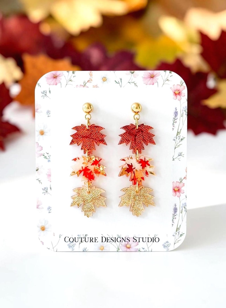 May include: Dangle earrings featuring three stacked maple leaf charms in red, white, and gold tones. The top leaf is textured red, the middle is white with red leaf patterns, and the bottom is textured gold. The earrings are displayed on a white card with floral accents and the text COUTURE DESIGNS STUDIO.