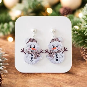 May include: A pair of snowman earrings. Each earring features a white snowman with a brown and white patterned hat and scarf. The snowman has black button eyes, an orange carrot nose, and a smiling mouth. The earrings have silver-colored posts.