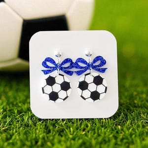 K&ouml;nnte beinhalten: Ohrringe mit Fu&szlig;ball-Designs in Schwarz und Wei&szlig;, verziert mit glitzernden blauen Schleifen. Pr&auml;sentiert auf einer wei&szlig;en Karte, vor dem Hintergrund von gr&uuml;nem Gras und einem Fu&szlig;ball.