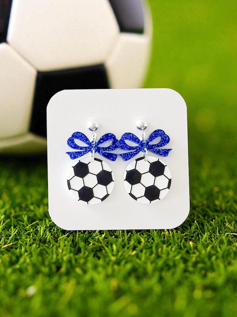 K&ouml;nnte beinhalten: Ohrringe mit Fu&szlig;ball-Designs in Schwarz und Wei&szlig;, verziert mit glitzernden blauen Schleifen. Pr&auml;sentiert auf einer wei&szlig;en Karte, vor dem Hintergrund von gr&uuml;nem Gras und einem Fu&szlig;ball.