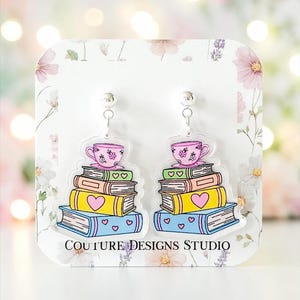 May include: A pair of dangle earrings featuring a stack of four colorful books topped with a pink teacup. The books are blue, yellow, peach, and green, decorated with heart motifs. The earrings are displayed on a white card labeled COUTURE DESIGNS STUDIO.