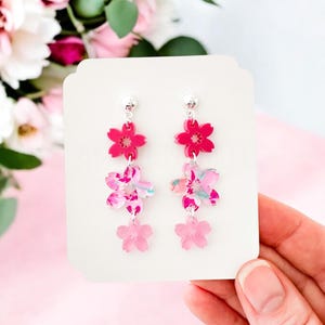 May include: A pair of pink and white floral dangle earrings with three flower charms on each earring. The earrings have silver posts and are displayed on a white card.