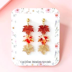 May include: Dangle earrings featuring three stacked maple leaf charms in red, white, and gold tones, suspended from gold-tone ball posts. The earrings are displayed on a white card with floral accents and the text COUTURE DESIGNS STUDIO.