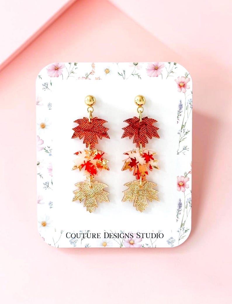 May include: Dangle earrings featuring three stacked maple leaf charms in red, white, and gold tones, suspended from gold-tone ball posts. The earrings are displayed on a white card with floral accents and the text COUTURE DESIGNS STUDIO.