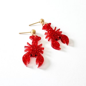 K&ouml;nnte beinhalten: Ein Paar rote Glitzer-Hummer-Ohrringe mit goldfarbenen Stiften und Kugelsteckern. Die Ohrringe sind mit kleinen schwarzen Augen versehen und vor einem wei&szlig;en Hintergrund platziert. Das Hummer-Design ist ein lustiges Accessoire mit nautischem Thema.