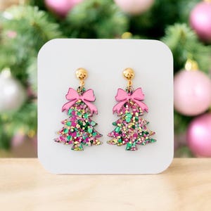 May include: A pair of Christmas tree earrings with pink bows and gold-colored studs. The earrings are shaped like Christmas trees and are filled with colorful glitter. The background features a white surface and a blurred Christmas tree.