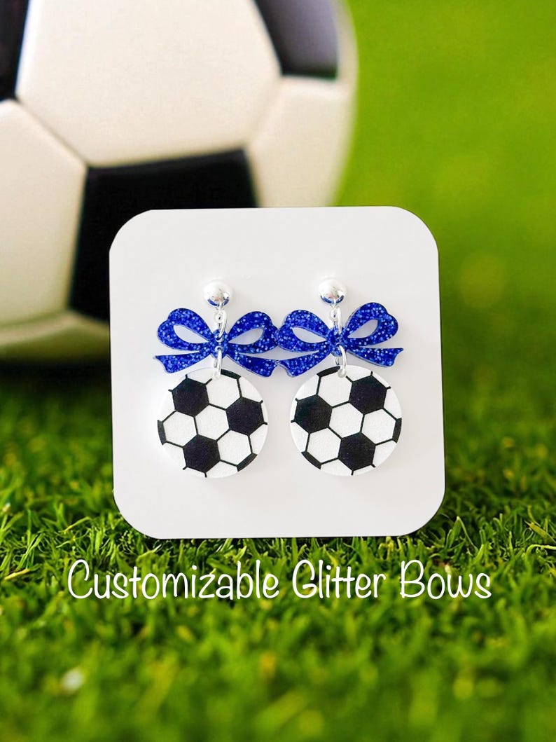 K&ouml;nnte beinhalten: Ohrringe mit einem schwarz-wei&szlig;en Fu&szlig;ball-Design, verziert mit blauen Glitzer-Schleifen. Die Ohrringe werden auf einer wei&szlig;en Karte mit dem Text "Customizable Glitter Bows" vor einem gr&uuml;nen Hintergrund und einem Fu&szlig;ball pr&auml;sentiert.