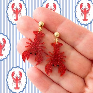K&ouml;nnte beinhalten: Ein Paar rote Glitzer-Hummer-Ohrringe mit goldfarbenen Kugelsteckern. Die Ohrringe werden vor einem blau-wei&szlig; gestreiften Hintergrund mit Hummer-Illustrationen gehalten. Ein lustiges Accessoire zum Thema Nautik.