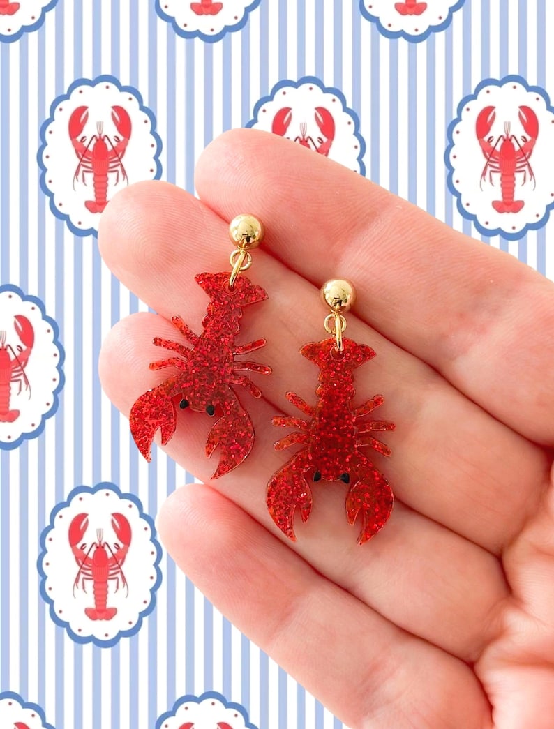 K&ouml;nnte beinhalten: Ein Paar rote Glitzer-Hummer-Ohrringe mit goldfarbenen Kugelsteckern. Die Ohrringe werden vor einem blau-wei&szlig; gestreiften Hintergrund mit Hummer-Illustrationen gehalten. Ein lustiges Accessoire zum Thema Nautik.