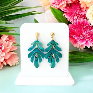 May include: Teal green leaf-shaped dangle earrings with gold accents. The earrings are displayed on a white stand against a light blue background.