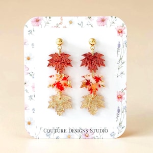 May include: Dangle earrings featuring three stacked maple leaf charms in metallic red, translucent white with red leaf patterns, and textured gold. The earrings have gold-tone stud posts and are displayed on a white card with floral print and the text COUTURE DESIGNS STUDIO.