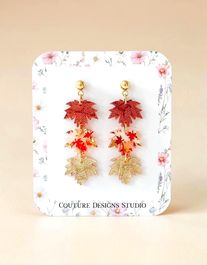 May include: Dangle earrings featuring three stacked maple leaf charms in metallic red, translucent white with red leaf patterns, and textured gold. The earrings have gold-tone stud posts and are displayed on a white card with floral print and the text COUTURE DESIGNS STUDIO.