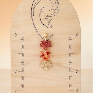 May include: A dangling earring featuring three stacked maple leaf charms in red, orange, and gold tones, suspended from a gold-tone ball stud. The earring is displayed on a wooden stand with an ear outline and a ruler marked in inches.