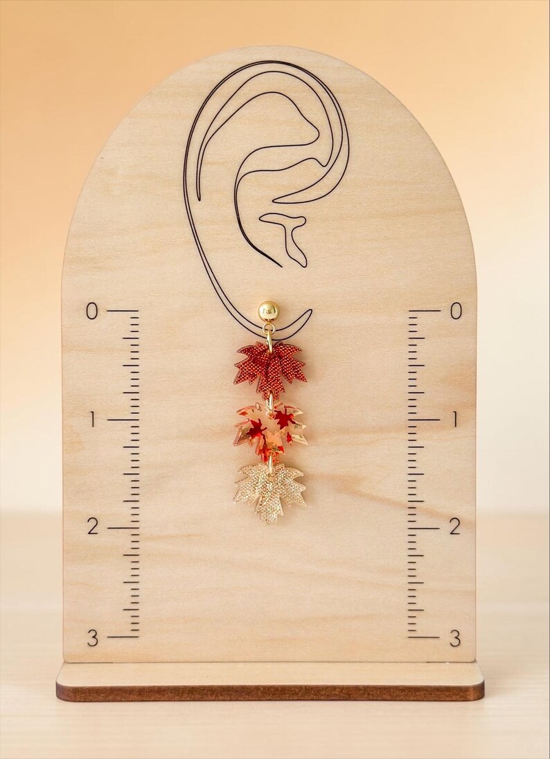 May include: A dangling earring featuring three stacked maple leaf charms in red, orange, and gold tones, suspended from a gold-tone ball stud. The earring is displayed on a wooden stand with an ear outline and a ruler marked in inches.