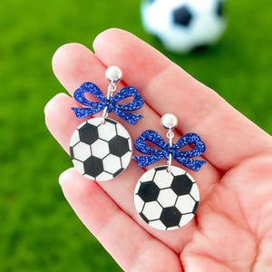 K&ouml;nnte beinhalten: Fu&szlig;ball-Ohrringe. Jeder Ohrring hat ein schwarz-wei&szlig;es Fu&szlig;ballmuster, das mit einer glitzernden blauen Schleife verziert ist. Die Ohrringe werden in einer Hand gehalten, mit einem unscharfen Fu&szlig;ball im Hintergrund.