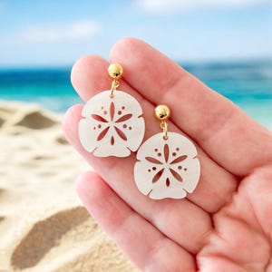 May include: A pair of white sand dollar earrings with gold-colored studs and hooks. The earrings are held in a hand against a backdrop of a sandy beach and ocean.