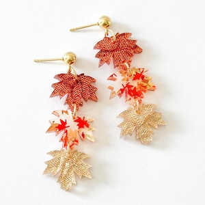 May include: A pair of dangle earrings featuring three stacked maple leaf charms in descending order. The top leaf is textured dark orange, the middle is translucent with red and orange patterns, and the bottom is textured gold. Gold-tone posts.
