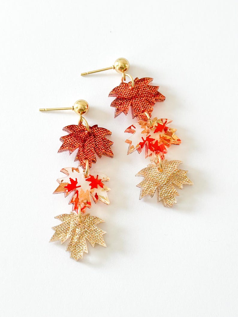 May include: A pair of dangle earrings featuring three stacked maple leaf charms in descending order. The top leaf is textured dark orange, the middle is translucent with red and orange patterns, and the bottom is textured gold. Gold-tone posts.