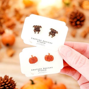 May include: Two pairs of stud earrings on white cards. The first pair features a brown glitter turkey design. The second pair features a pumpkin design with orange glitter. Both cards have the text "Couture Designs Studio" printed on them.