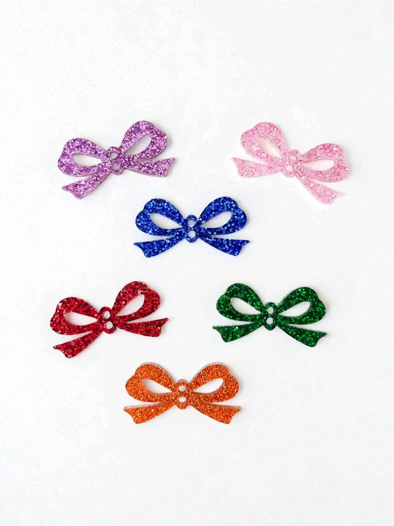 K&ouml;nnte beinhalten: Sechs glitzernde Schleifen in verschiedenen Farben: Lila, Rosa, Blau, Rot, Gr&uuml;n und Orange. Diese dekorativen Schleifen sind auf wei&szlig;em Hintergrund angeordnet und zeigen ihre glitzernde Textur und lebendigen Farbt&ouml;ne.