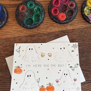 Puede incluir: Una tarjeta de felicitaci&oacute;n rectangular con cuatro fantasmas dibujados a mano que sostienen varias bebidas alcoh&oacute;licas, calabazas naranjas y una telara&ntilde;a. El texto I'M HERE FOR THE BOOS! est&aacute; escrito en tinta negra en el centro, rodeado de peque&ntilde;os puntos naranjas y negros.