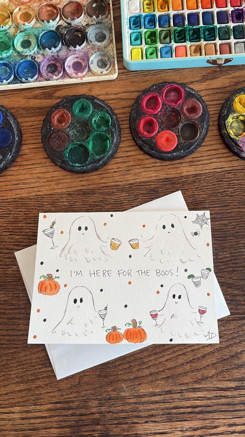 Puede incluir: Una tarjeta de felicitaci&oacute;n rectangular con cuatro fantasmas dibujados a mano que sostienen varias bebidas alcoh&oacute;licas, calabazas naranjas y una telara&ntilde;a. El texto I'M HERE FOR THE BOOS! est&aacute; escrito en tinta negra en el centro, rodeado de peque&ntilde;os puntos naranjas y negros.