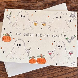 Puede incluir: Una tarjeta de felicitaci&oacute;n rectangular con fondo blanco que presenta cuatro fantasmas dibujados a mano sosteniendo varias bebidas alcoh&oacute;licas. El texto "I'M HERE FOR THE BOOS!" est&aacute; centrado. Se ilustran calabazas naranjas, puntos negros y naranjas, una ara&ntilde;a y una telara&ntilde;a.