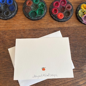 Puede incluir: Una tarjeta de felicitaci&oacute;n rectangular blanca plegada con una peque&ntilde;a ilustraci&oacute;n de una calabaza naranja y el texto Amy and Alfred Designs impreso en tinta negra en la parte inferior central, sobre un sobre blanco a juego.