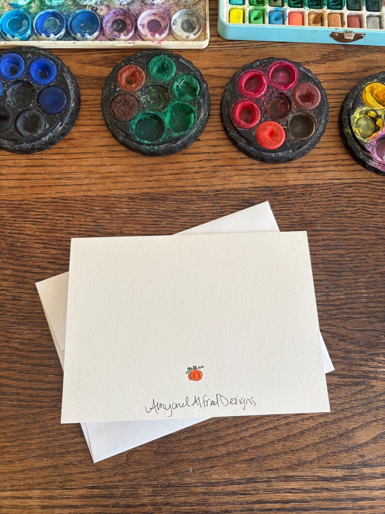 Puede incluir: Una tarjeta de felicitaci&oacute;n rectangular blanca plegada con una peque&ntilde;a ilustraci&oacute;n de una calabaza naranja y el texto Amy and Alfred Designs impreso en tinta negra en la parte inferior central, sobre un sobre blanco a juego.