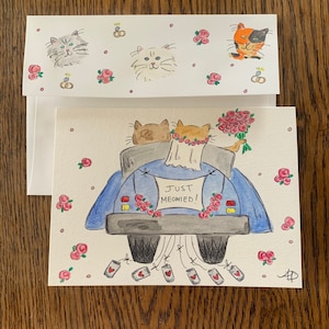 May include: A wedding card featuring a blue car with two cats inside. The car has a sign that says "Just Meownied!" and is decorated with red roses and cans tied to the back. The card is decorated with a white background and pink roses.