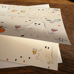 Puede incluir: Una tarjeta de felicitaci&oacute;n blanca con sobre a juego que presenta ilustraciones dibujadas a mano de fantasmas sosteniendo varias bebidas alcoh&oacute;licas. La tarjeta incluye el texto "HERE FOR THE BOOS!" junto con peque&ntilde;os puntos naranjas y negros, una telara&ntilde;a y una calabaza.