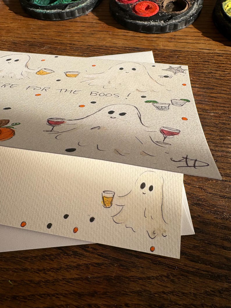 Puede incluir: Una tarjeta de felicitaci&oacute;n blanca con sobre a juego que presenta ilustraciones dibujadas a mano de fantasmas sosteniendo varias bebidas alcoh&oacute;licas. La tarjeta incluye el texto "HERE FOR THE BOOS!" junto con peque&ntilde;os puntos naranjas y negros, una telara&ntilde;a y una calabaza.