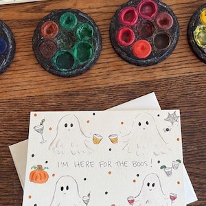 Puede incluir: Una tarjeta de felicitaci&oacute;n rectangular con cuatro fantasmas dibujados a mano que sostienen varios c&oacute;cteles, calabazas naranjas y puntos negros y naranjas. El texto "I'M HERE FOR THE BOOS!" est&aacute; escrito con tinta negra en el centro.