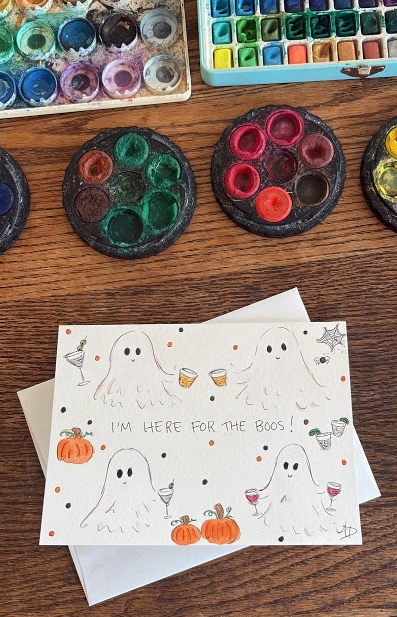 Puede incluir: Una tarjeta de felicitaci&oacute;n rectangular con cuatro fantasmas dibujados a mano que sostienen varios c&oacute;cteles, calabazas naranjas y puntos negros y naranjas. El texto "I'M HERE FOR THE BOOS!" est&aacute; escrito con tinta negra en el centro.
