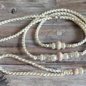 Op de afbeelding: Een witte gevlochten touwlijn met parel- en steenaccenten. De lijn heeft een zilveren sluiting en een klein rond label met tekst.