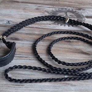 Könnte beinhalten: Ein schwarzes geflochtenes Band mit silbernen Metall-Akzenten und einem schwarzen Leder-Armband. Das Band ist auf einer Holzoberfläche geschlungen und aufgerollt.