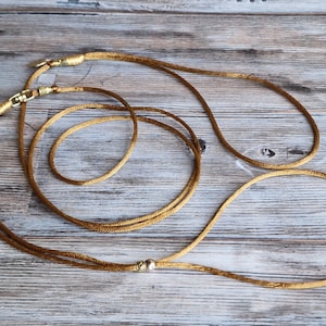 May include: A long, thin, golden-brown cord necklace featuring a smooth, metallic silver-toned bead accent. The cord is finished with gold-toned metal end caps and a lobster claw clasp, creating a simple, continuous loop design for jewelry.