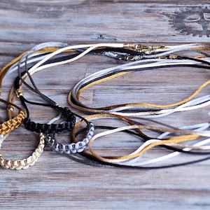 Può includere: Una collezione di diverse collane con cordini sottili in oro metallizzato, argento, nero e bianco. Ogni collana include una sezione intrecciata e un piccolo ciondolo circolare in metallo. I cordini sono disposti in anelli sovrapposti con chiusure.