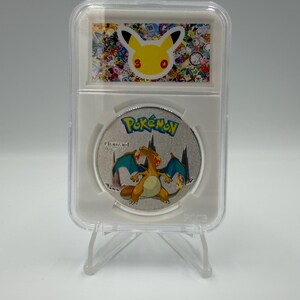 May include: A silver-colored circular collectible coin featuring a colorful Charizard character illustration and the Pokémon logo, encased in a clear rectangular protective plastic slab with a Pikachu 30th anniversary graphic label at the top.